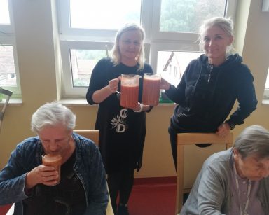 Kierowniczka Ilona Gajewska, neurologopedka Anna Szmaja-Wysocka trzymają dzbanki z sokami. Przed nimi seniorki piją soki.