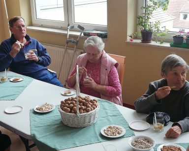 Troje seniorów przy stole. Jedzą deser ze szklanek. przed nimi na blacie kosz orzechów, miseczki z orzechami i nasionami.