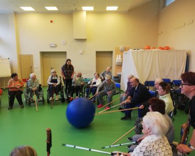 Seniorzy i seniorzy siedzą w kręgu na sali gimnastycznej. Drewnianymi kijkami odpychają niebiską piłkę.