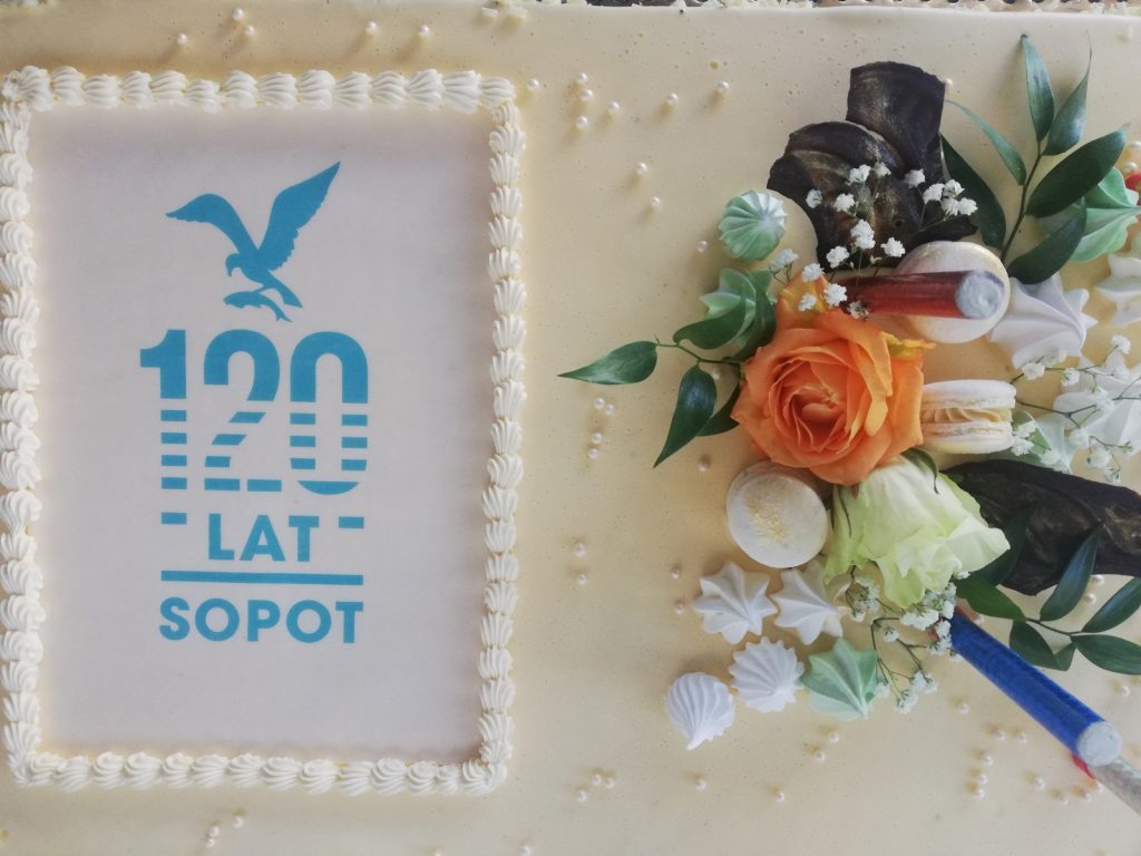 Biały tort. Na nim dekoracja z kwiatów, pereł, ciasteczek, napis "120 lat Sopot", obrazek mewy trzymającej rybę.