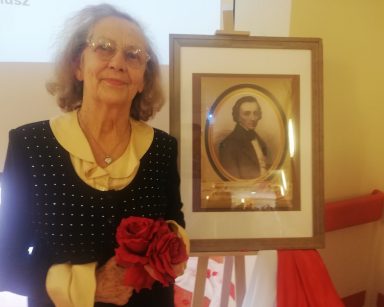 Seniorka pozuje przy portrecie Fryderyka Chopina. Trzyma w dłoniach czerwone róże.