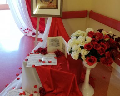 Na stelażu portret Fryderyka Chopina w ramie. Dekoracja. Na białym i czerwonym materiale kartki z nutami. Białe i czerwone kwiaty.