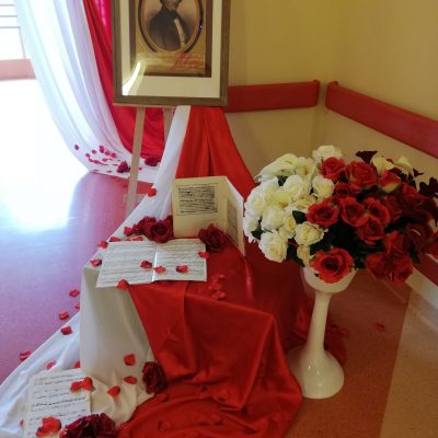 Na stelażu portret Fryderyka Chopina w ramie. Dekoracja. Na białym i czerwonym materiale kartki z nutami. Białe i czerwone kwiaty.