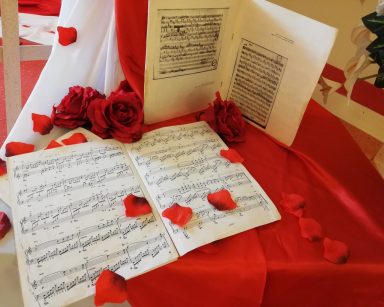 Dekoracja. Na białym i czerwonym materiale kartki z nutami, płatki czerwonych róż, czerwone róże.