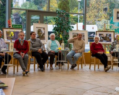 Przy stolikach w ogrodzie zimowym siedzą seniorzy. Ogród udekorowany oprawionymi w ramy zdjęciami Sopotu.