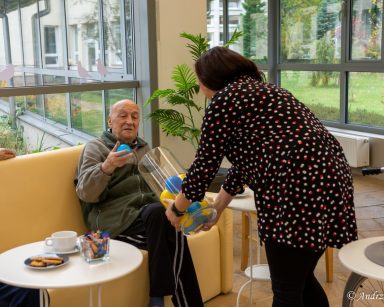 Terapeutka Małgorzata Jancelewicz trzyma naczynie z kolorowymi kulami. Senior wyciąga niebieską kulę.