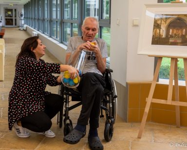 Terapeutka Małgorzata Jancelewicz trzyma naczynie z kolorowymi kulami. Senior wyciąga żółtą kulę.