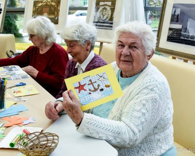 Warsztaty plastyczne. Seniorka pokazuje wykonaną metodą kolażu pocztówkę z napisem Sopot. Obok siedzą dwie seniorki.
