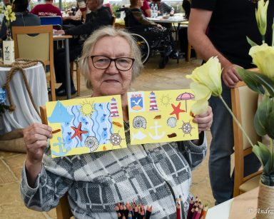 Seniorka prezentuje dwie pocztówki wykonane techniką kolażu. Na nich motywy kojarzące się z Sopotem i morzem.