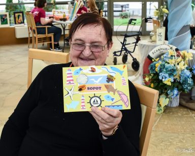 Seniorka prezentuje pocztówkę. Na niej naklejony napis 