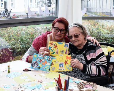 Warsztaty plastyczne. Koordynatorka Edyta Życzyńska i seniorka prezentują wykonane pocztówki. Obejmują się i uśmiechają.