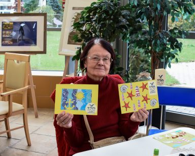 Ogród zimowy. Uśmiechnięta seniorka pokazuje dwie pocztówki wykonane na 120. urodziny miasta Sopotu.