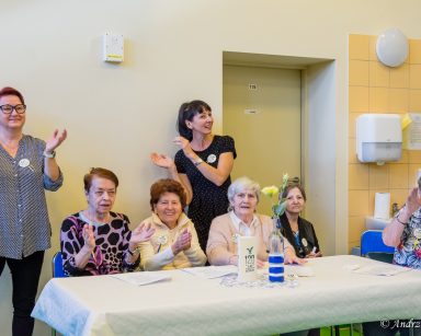 Seniorki siedzą przy stole śmieją się i klaszczą. Za nimi terapeutka Ania Rzepczyńska i koordynatorka Edyta Życzyńska.