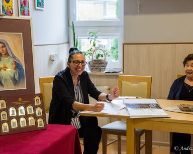 Przy stole terapeutka Magdalena Poraj-Górska i seniorka. Obok obraz Matki Boskiej i sztabki z małymi portretami Maryi.