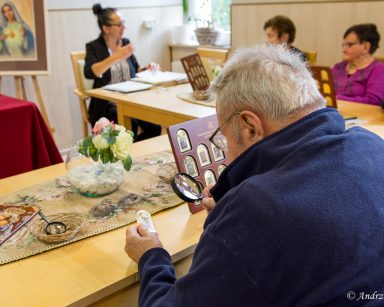 Terapeutka Magdalena Poraj-Górska i seniorzy przy stole. Senior ogląda przez lupę obraz Matki Boskiej na złotej sztabce.