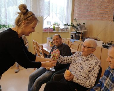 Trzech uśmiechniętych seniorów siedzi obok siebie. Terapeutka Beata Brzozowska nachyla się i częstuje ich lizakami.