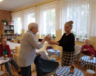 Terapeutka Beata Brzozowska częstuje seniorkę lizakiem. Obok przy stolikach siedzą dwie seniorki.