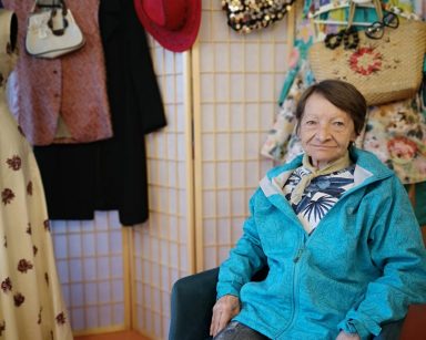 Uśmiechnięta seniorka na krześle. Za nią parawan i kolorowe ubrania.