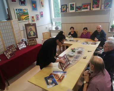 Sala. Terapeutka Magdalena Poraj-Górska pokazuje seniorom tablice z kolekcją obrazów Matki Boskiej na złotych sztabkach.
