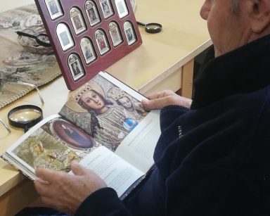 Senior ogląda książkę z obrazami Maryi. Na stole obrazy Matki Boskiej na złotych sztabkach. Sztabki są w drewnianych ramach.