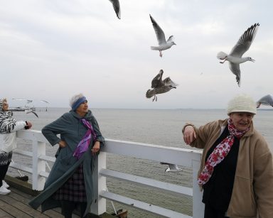 Trzy seniorki na molo w Sopocie. Koło nich latają mewy. Za barierką morze.