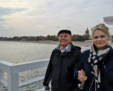 Molo w Sopocie. Terapeutka Beata Brzozowska i senior spacerują za rękę. Uśmiechają się.