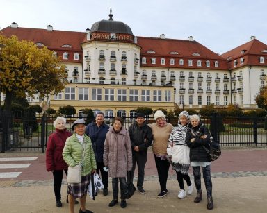 Grupa seniorów pozuje do zdjęcia. Mają ciepłe kurtki. Uśmiechają się. Za nimi Grand Hotel w Sopocie.