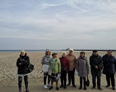 Grupa seniorów pozuje do zdjęcia. Mają ciepłe kurtki. Za nimi plaża, morze, molo.