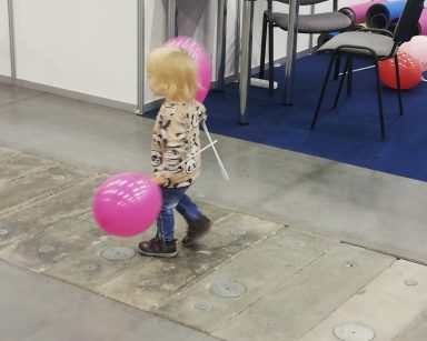 Hala. Przy stoisku małe dziecko. W obu dłoniach trzyma różowe balony na patyku.