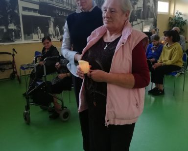 Sala. Terapeutka Magdalena Poraj-Gorska i seniorka idą złożyć świeczkę. Za nimi seniorzy siedzą w rzędach.