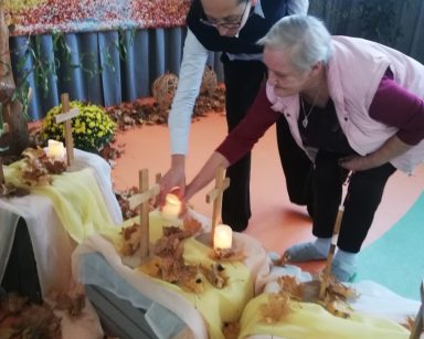 Sala z jesienną dekoracją. Terapeutka Magdalena Poraj-Górska i seniorka składają świeczkę pod krzyżem.