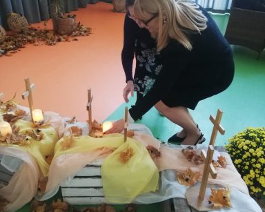 Sala z jesienną dekoracją. Dyrektorka Agnieszka Cysewska i seniorka składają świeczkę pod krzyżem.