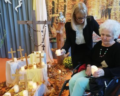 Sala z jesienną dekoracją. Dyrektorka Agnieszka Cysewska i seniorka składają świeczki wśród drewnianych krzyży.