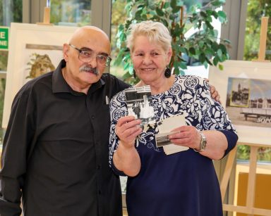 Andrzej Zięba obejmuje ręka seniorkę. Seniorka pokazuje biało-czarna fotografię. Obydwoje się uśmiechają.