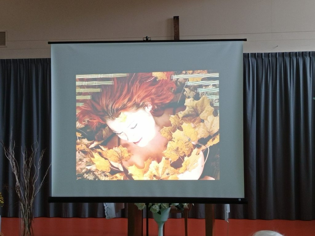 Sala. Ekran projektora. Kobieta z rudymi włosami. Jest obsypana żółtymi, jesiennymi liści.