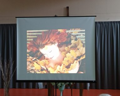Sala. Ekran projektora. Kobieta z rudymi włosami. Jest obsypana żółtymi, jesiennymi liści.