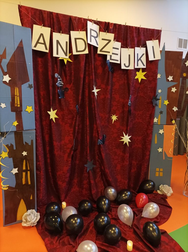 Sala. Andrzejkowa dekoracja. Napis "Andrzejki", czerwona zasłona z gwiazdami, nietoperzami, kotami, na podłodze balony.