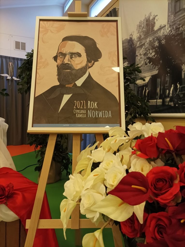 Na stelażu portret mężczyzny. Napis "2021 rokiem Cypriana Kamila Norwida". Obok bukiet białych i czerwonych kwiatów.