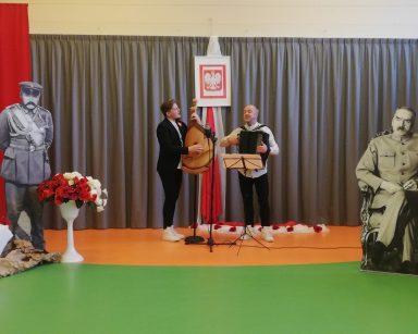 Sala z dekoracją z okazji Święta Niepodległości. Dwaj muzycy z zespołu Galicia Folk Band. Grają i śpiewają.