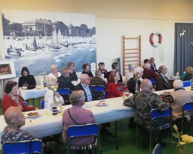 Sala. Spotkanie Związku Emerytów, Rencistów i Inwalidów. Seniorzy i goście przy stołach, rozmawiają.