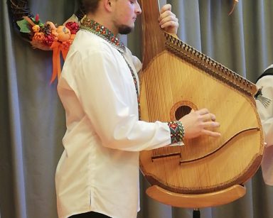 Muzyk gra na ukraińskim instrumencie - bandurze. Ma białą koszulę z mankietami i kołnierzem w ludowym wzorze.