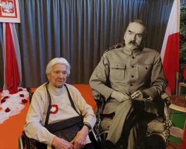 Sala. Przy tekturowej postaci Piłsudskiego siedzi seniorka. Dekoracja z okazji Święta Niepodległości.
