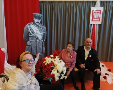 Sala. Przy tekturowej postaci Piłsudskiego siedzi troje seniorów. Dekoracja z okazji Święta Niepodległości.