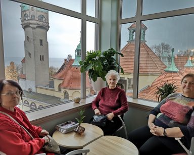 Kawiarnia. Trzy seniorki przy stole. Za oknem dachy budynków, morze.