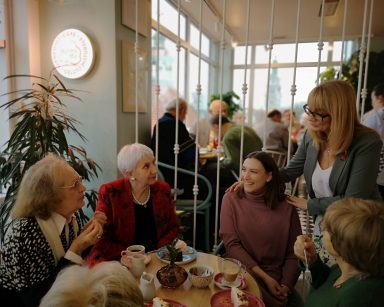 Kawiarnia. Seniorzy, pracownicy, dyrektorka Cysewska. Siedzą przy stolikach, stoją, piją kawę, jedzą ciasto, rozmawiają.