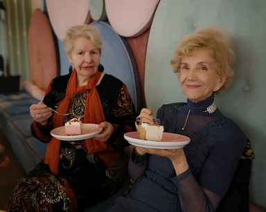 Kawiarnia. Dwie odświętnie ubrane seniorki siedzą na kanapie. Trzymają talerzyki z ciastem, uśmiechają się.