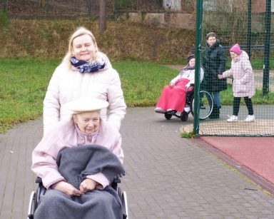 Seniorka na wózku inwalidzkim, za nią neurologopedka Anna Szmaja-Wysocka. Są ciepło ubrane. Dalej spacerowicze, trawnik, las.