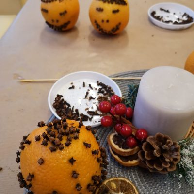 Na blacie stołu świąteczne ozdoby. Pomarańcze udekorowane goździkami, szyszki, plastry suszonych pomarańczy, świeczka.