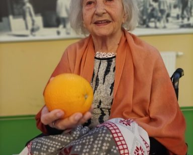 Sala. Seniorka siedzi na wózku. Uśmiecha się. Nogi ma okryte kocem. Wyciąga rękę, w dłoni trzyma pomarańczę.