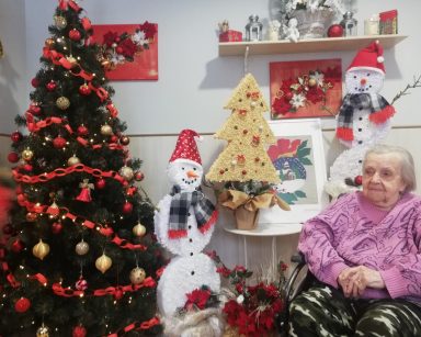 Sala ze świąteczną dekoracją. W rogu przystrojona choinka. Świąteczne figury: dwa bałwanki, choinka. Obok siedzi seniorka.